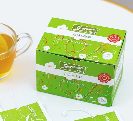 Green tea box with floral design and tea bag next to a glass cup of green tea on white surface